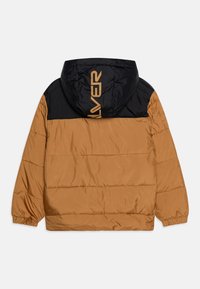 Doudoune rembourrée avec une capuche noire et une partie supérieure noire, ainsi qu'une partie inférieure beige. Comprend des poignets élastiques et un logo orange au dos.