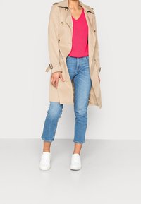 Beige trenchcoat met gouden knopen, gedragen over een roze geribbelde trui. Gecombineerd met lichtblauwe, enkelhoge jeans en witte sneakers.