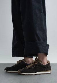 Inimene, kes kannab tumepruune Michael Korsi seemisnahast tossusid, millel on tanvärvi paelad ja ääris, kombineeritud laia musta mansetiga pükstega hallil põrandal.