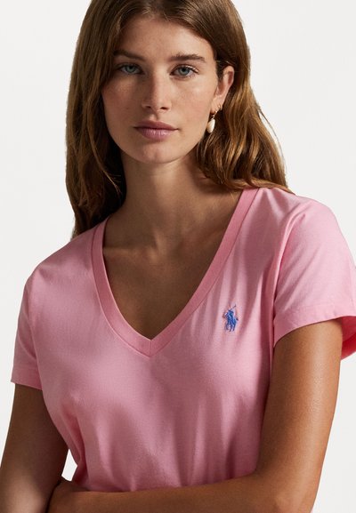 T-shirt en coton rose avec un design à col en V, des manches courtes et un petit logo brodé bleu sur la poitrine gauche. Texture lisse.