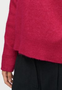 Pull en tricot fuchsia à texture douce, avec des poignets côtelés et un ourlet court, assorti à un pantalon noir. Aucun motif ou accent visible.