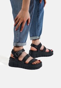 Sandali con platform neri e suola spessa, dotati di cinturini larghi e dettaglio con fibbia. Indossati con jeans blu arrotolati.
