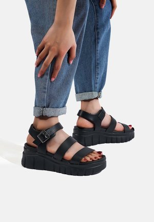 Sandali con platform neri e suola spessa, dotati di cinturini larghi e dettaglio con fibbia. Indossati con jeans blu arrotolati.