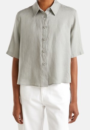 Camisa de manga corta gris claro con botones y cuello puntiagudo, usada con pantalones blancos en una persona de pie.