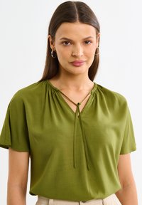 Žalia palaidinė trumpomis rankovėmis, V formos iškirpte, raukšlėmis ir reguliuojamomis raištelėmis iškirptėje. Lygus audinys su subtiliu blizgesiu.