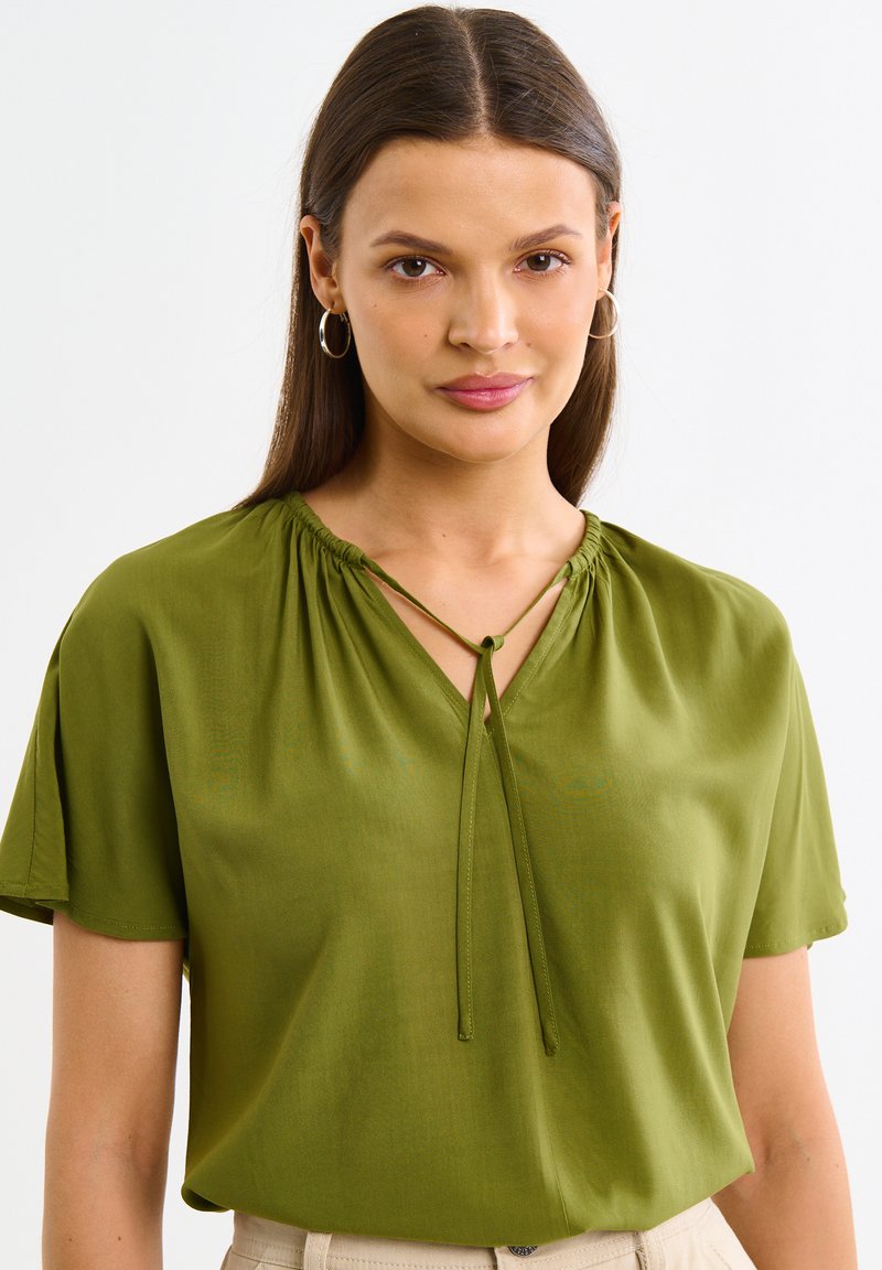 Roheline pluus lühikeste varrukate, V-kaeluse, kogutud detaili ja reguleeritavate paeltega kaeluses. Sile kude, millel on peen sära.