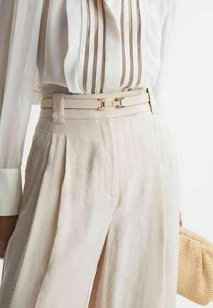 Crème-kleurige high-waisted broeken met plooien en zijzakken, gecombineerd met een doorzichtige witte blouse met verticale bruine strepen.