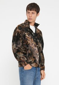 Jachetă din fleece cu fermoar, având un model multicolor de frunze în nuanțe de maro și negru. Guler înalt, manșete elastice și buzunare laterale.