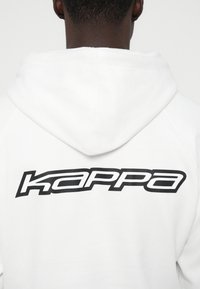 Rückansicht einer Person in einem weißen Hoodie mit hochgezogenem Kapuzen und dem schwarzen "Kappa"-Logo, das sich über den oberen Rücken erstreckt.