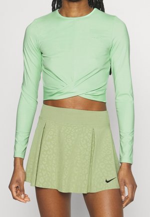 Kvinde iført en lysegrøn langærmet crop top og en lysegrøn plisseret sportnederdel med sort Nike-logo på kanten.