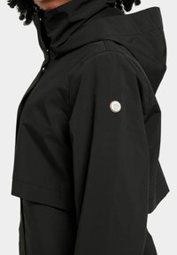 Veste imperméable noire avec un col haut, présentant un logo circulaire contrasté sur la manche gauche et un design élégant et minimaliste.