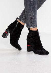 Schwarze, samtartige Knöchelschuhe mit spitzer Zehe und Reißverschluss. Sie verfügen über einen klobigen, glänzenden, schwarzen Absatz mit einem roten Akzentband.