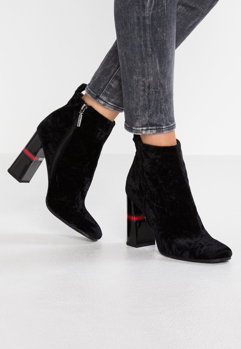 Schwarze, samtartige Knöchelschuhe mit spitzer Zehe und Reißverschluss. Sie verfügen über einen klobigen, glänzenden, schwarzen Absatz mit einem roten Akzentband.