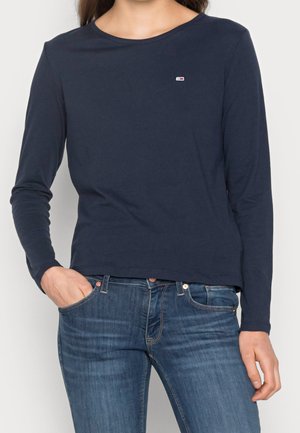 Personne portant une chemise bleu marine à manches longues avec un petit logo sur la poitrine et un jean bleu, une main dans la poche.