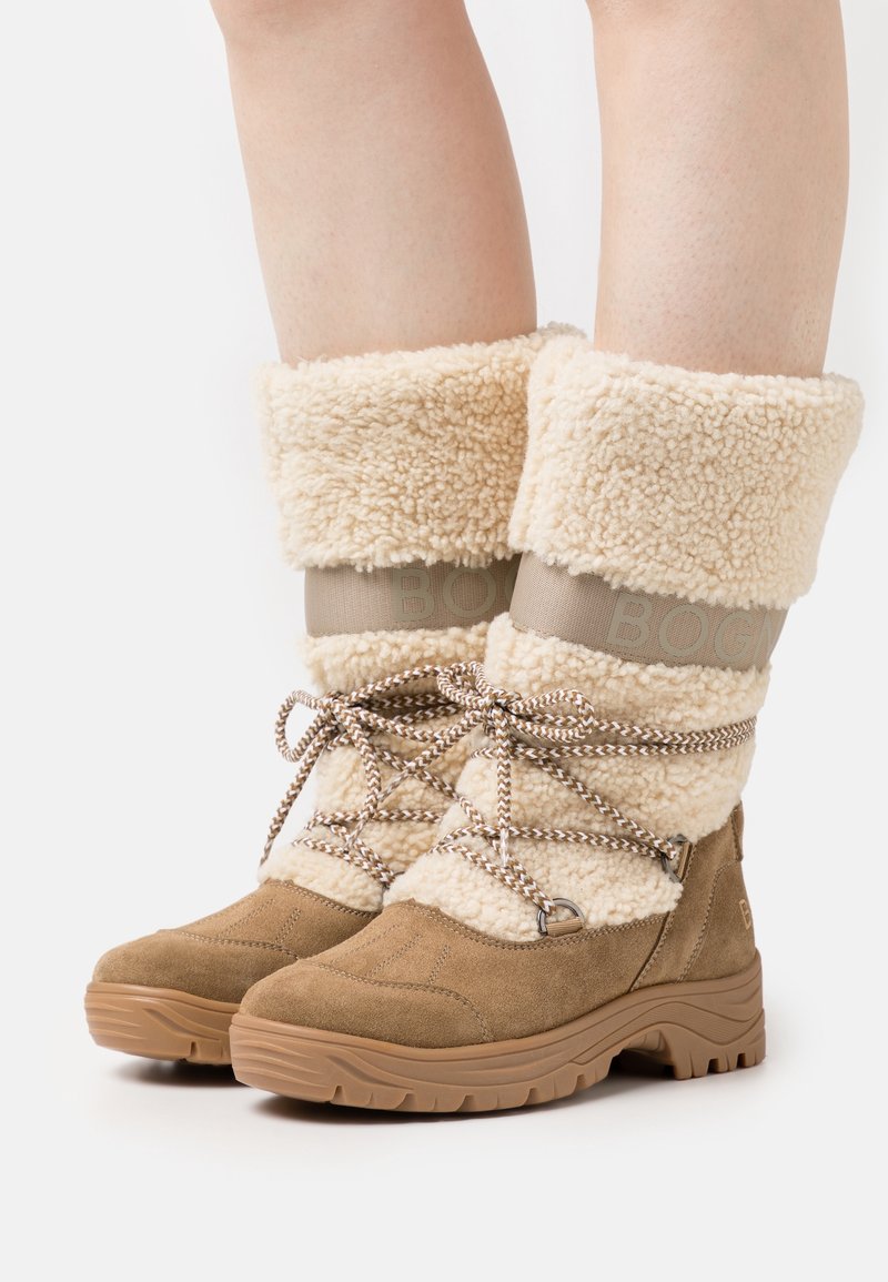 Botas de ante color arena con un puño de borrego esponjoso en color crema. Presentan una banda con logotipo beige, cordones trenzados y suela de goma texturizada para un mejor agarre.