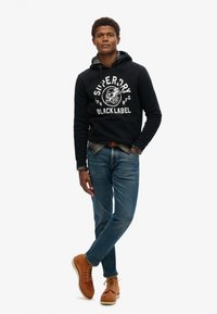 Sudadera negra con texto gráfico blanco, jeans de mezclilla azul y botas marrones con cordones. Camisa de franela notable debajo de la sudadera. Estilo casual.