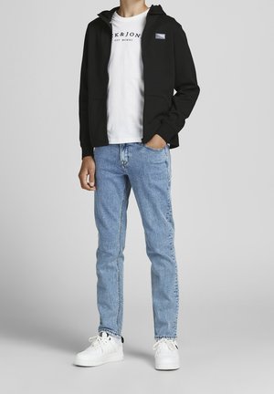 Person med ljusblå jeans, vita sneakers, vit grafisk t-shirt och svart zip-up hoodie med händerna i fickorna mot en grå bakgrund.