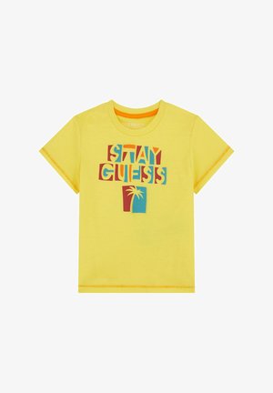 T-shirt di cotone giallo con grafica testuale multicolore "STAY GUESS" e una palma. Maniche corte con dettagli di cucitura a contrasto.