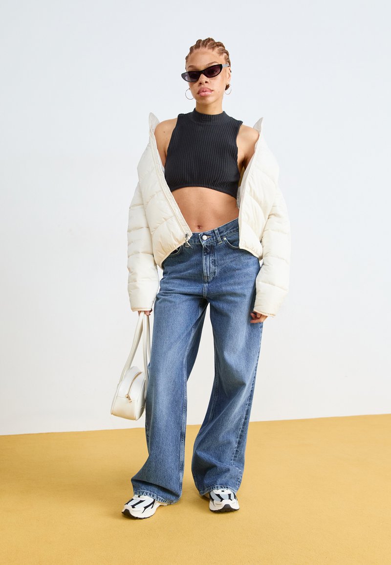 Kurtka puchowa w kolorze off-white, prążkowany czarny top crop, szerokie niebieskie dżinsy, czarno-białe trampki, trzymając małą białą torebkę.
