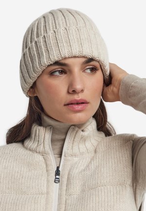 Kobieta w beżowej prążkowanej dzianej czapce i pasującym swetrze na zamek, patrząca w bok, jedną ręką dotykająca głowy.