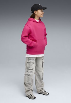 Ung person iført lyserød hættetrøje, lysegrå cargobukser, sort kasket og grå sneakers, vendt mod højre med hænderne i hættetrøjens lomme.