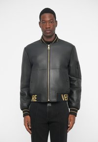 Νεαρός άνδρας φορόντας μια μαύρη δερμάτινη bomber jacket με χρυσές ρίγες στον γιακά και τα μανίκια, στέκεται μπροστά σε λευκό φόντο.