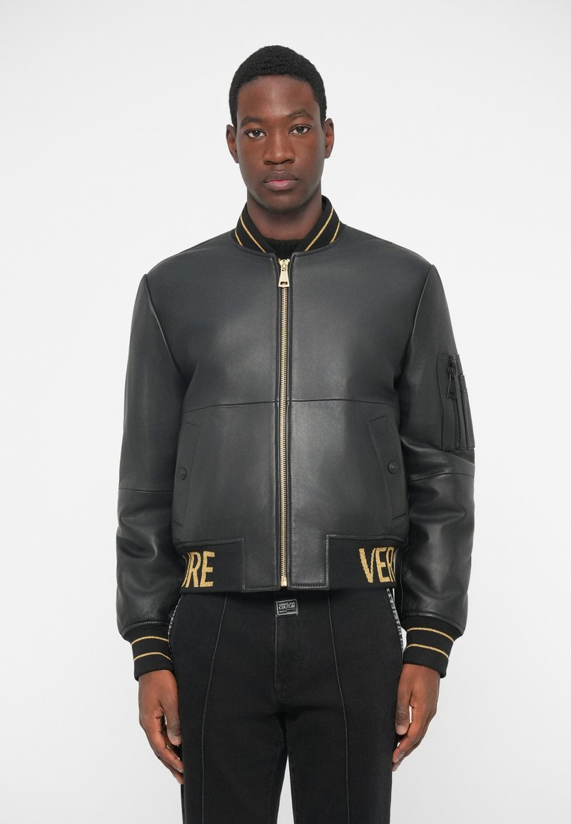 Νεαρός άνδρας φορόντας μια μαύρη δερμάτινη bomber jacket με χρυσές ρίγες στον γιακά και τα μανίκια, στέκεται μπροστά σε λευκό φόντο.