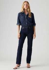 Donkerblauwe denimoverhemd met knoopsluiting, twee borstzakken en opgerolde mouwen, gecombineerd met high-waisted donkerblauwe Jeans en zwarte haksandalen.