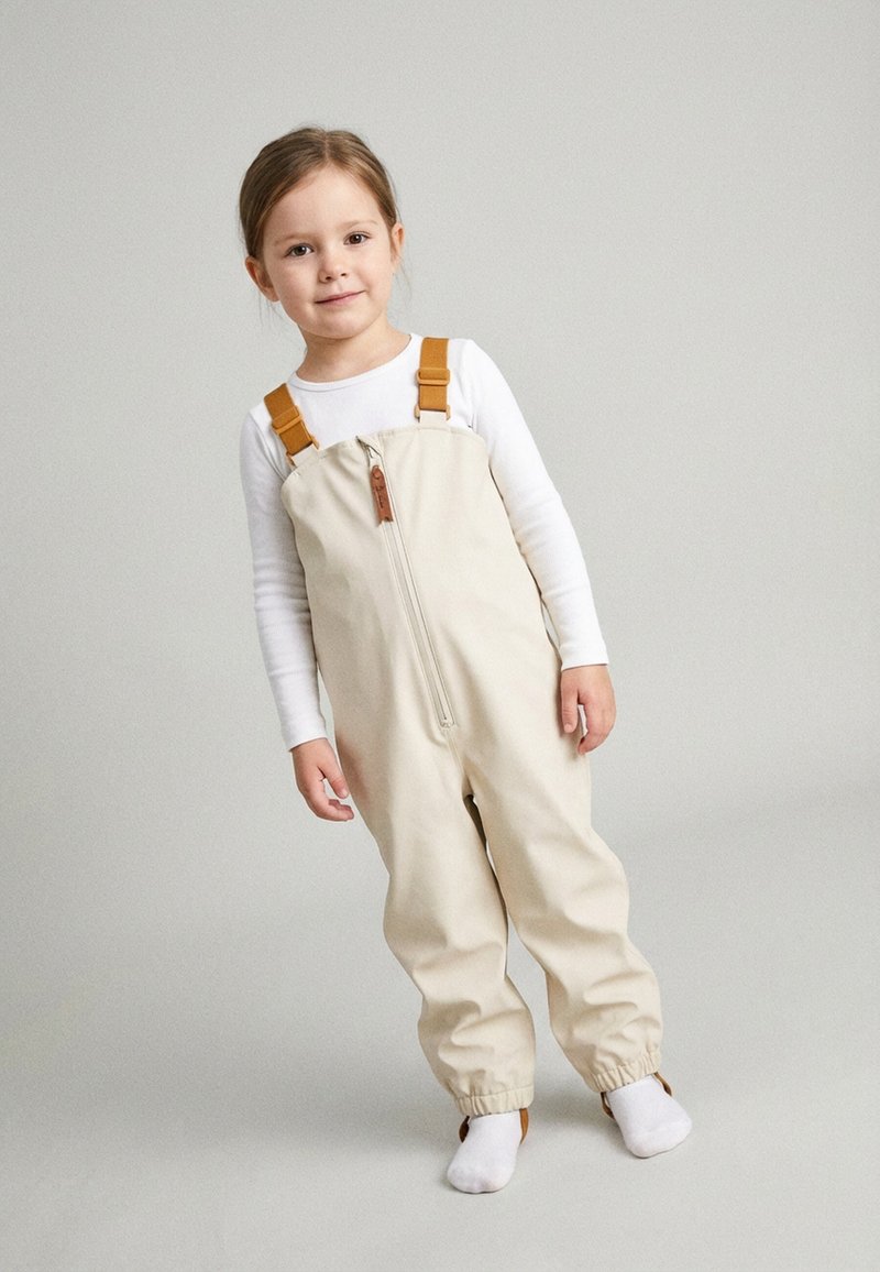 Junges Mädchen steht, trägt beige Latzhosen mit orangen Trägern über einem weißen Langarmshirt und weißen Socken, vor einem einfarbigen grauen Hintergrund.