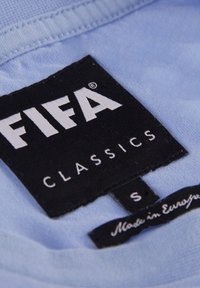 Étiquette de vêtement bleu clair avec une étiquette rectangulaire noire affichant "FIFA CLASSICS" en texte blanc gras, taille "S" et le détail "Fabriqué en Europe".
