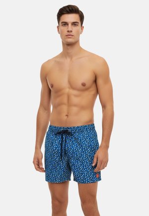 Uomo senza maglietta con capelli corti castani che indossa pantaloncini da bagno blu con motivo bianco e giallo, in piedi davanti a uno sfondo semplice.