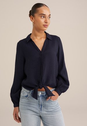 Navyblaue, leichte Button-up-Bluse mit langen Ärmeln und einem Band in der Taille. Texturiert, mit lässiger Passform, kombiniert mit hellblauen Jeans.