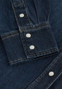 Țesătura de denim închis prezintă un tiv împăturit cu trei butoane cu presiune albe și cusături contrastante portocalii de-a lungul marginii.