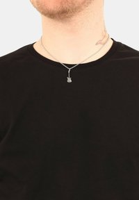 Collana in argento con un piccolo ciondolo a forma di chitarra, indossata sopra una t-shirt nera a girocollo. Il ciondolo presenta dettagli intricati.