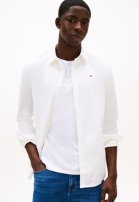 Camisa branca de manga comprida com colarinho abotoado, usada sobre uma t-shirt branca. Pequeno logótipo notável no peito. Corte casual com mangas enroladas.