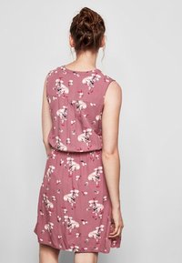 Kvinna med flätat hår klädd i en ärmlös, knälång klänning i dammig rosafärg med vita och rosa blommönster, stående mot en enkel bakgrund.