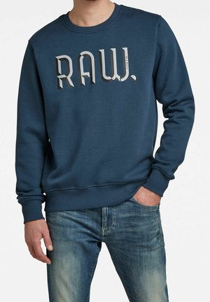Man draagt een marineblauwe trui met de tekst "RAW." en blauwe vervaagde jeans, staat met één hand in de zak en de andere ontspannen langs het lichaam.