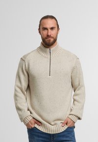 Beige striktrøje med en ribbet høj krave, halv lynlås lukning og tekstureret mønster overalt. Lange ærmer med ribbede manchetter.