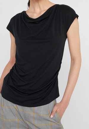 Mujer con una blusa negra de manga corta con caída y pantalones gris a cuadros con rayas amarillas, mano apoyada en el bolsillo del pantalón.