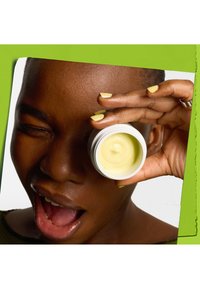 Weißes Glas mit gelber Creme, gehalten von einer Person. Der Hintergrund hat leuchtend grüne Akzente. Die Person hat kurze Haare und gelb lackierte Nägel.