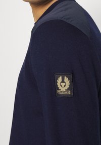 Námořnický svetr s texturovaným pleteným tělem, hladkými látkovými akcenty na ramenou a logem na levé paži. Jednoduchý, moderní design.