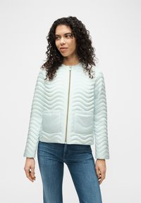 Γυναίκα με σγουρά μαλλιά που φοράει ανοιχτό μπλε καπιτονέ μπουφάν με εμπρόσθιες τσέπες και φερμουάρ, σε συνδυασμό με μπλε τζιν, πάνω σε απλό φόντο.