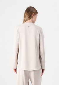 Blusa beige a maniche lunghe con una texture liscia, scollatura tonda e un dettaglio con bottone di accentuazione sul retro. Fit morbido e sartoriale.