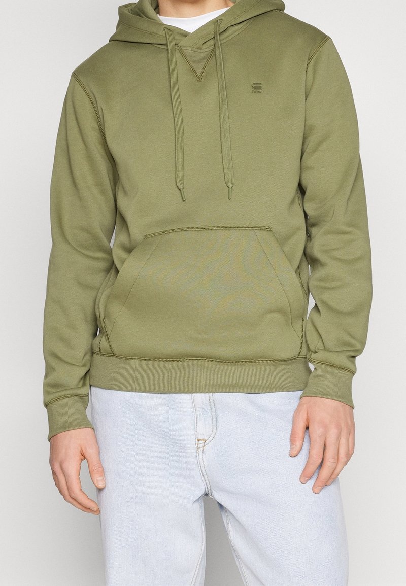 Personne portant un sweat à capuche vert olive avec poche avant et un jean bleu clair, montré du cou aux hanches.