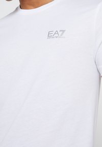 Bílé tričko s krátkými rukávy z měkké bavlny. Na hrudi má malý stříbrný vyšívaný logo EA7. Jednoduchý střih s kulatým výstřihem.