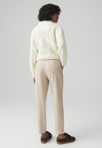 Crème kabelgebreide trui, beige op maat gemaakte broek en bruine sneakers. De trui heeft een hoge kraag, terwijl de broek een achterzak heeft.