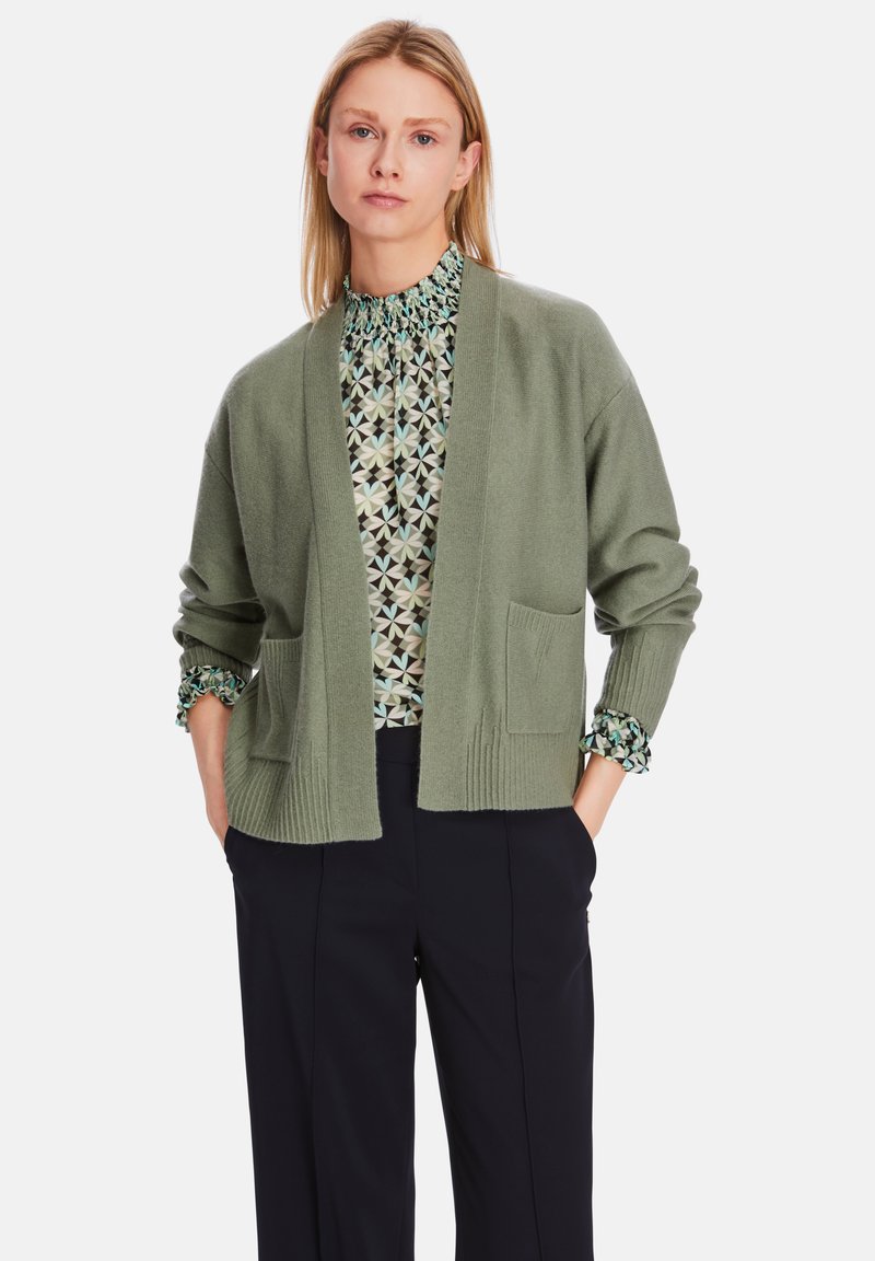 Olivgrüner Cardigan mit offenem Vorderteil, gerippter Textur und zwei Vordertaschen, getragen über einer gemusterten Bluse und kombiniert mit dunklen Hosen.