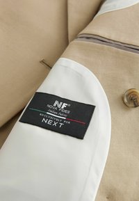 Tessuto di giacca beige con una fodera bianca liscia, caratterizzato da un'etichetta nera con la dicitura "NF NOVA FIDES Tessuto Italiano" e il testo "Esclusivamente per NEXT".