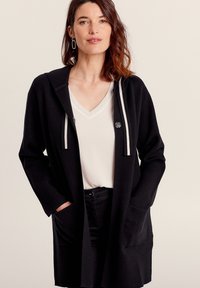 Cardigan nero con cappuccio e due tasche frontali, dotato di cordini grigi a righe e chiusura con un singolo bottone. Tessuto morbido e testurizzato.