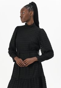 Abito lungo nero in tessuto testurizzato con maniche lunghe, scollatura alta, vita corsetto e gonna a strati; presenta dettagli in pizzo sulle maniche e motivi intricati.